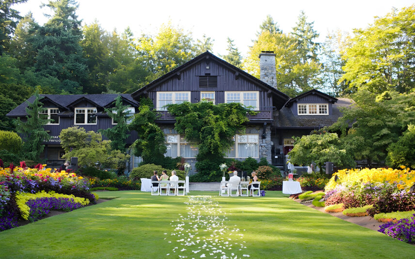 A small ceremony is beginning in front of the Stanley Park Pavilion, on a summer's day, petals lining the lawn
