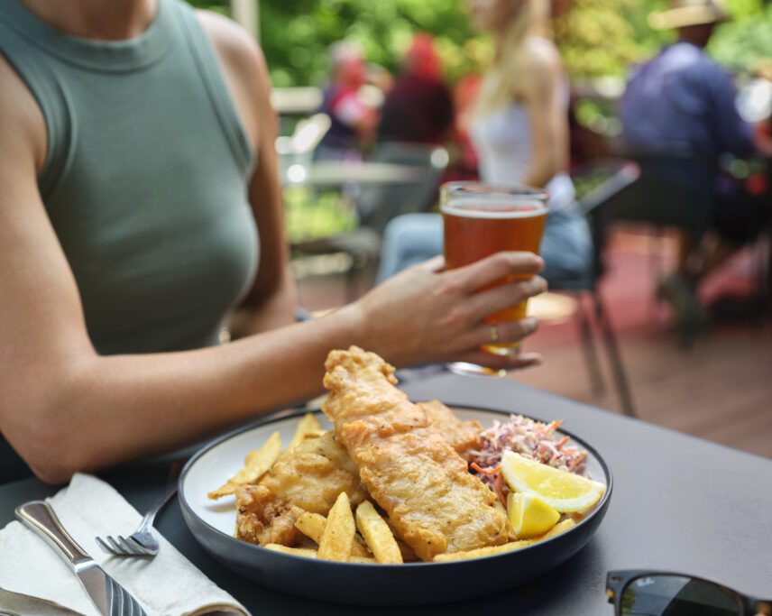 A woman holding a beer, while sat on the patio of Stanley's Bar and Grill. In front of her sits a huge plate of fish and chips.