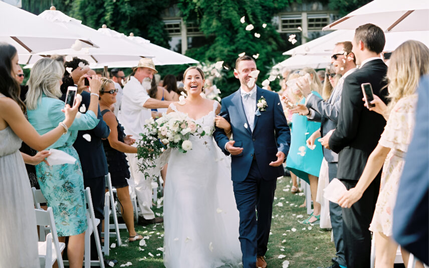 A couple walking down the aisle following their wedding ceremony, petals are in the air as folks applaud and smile