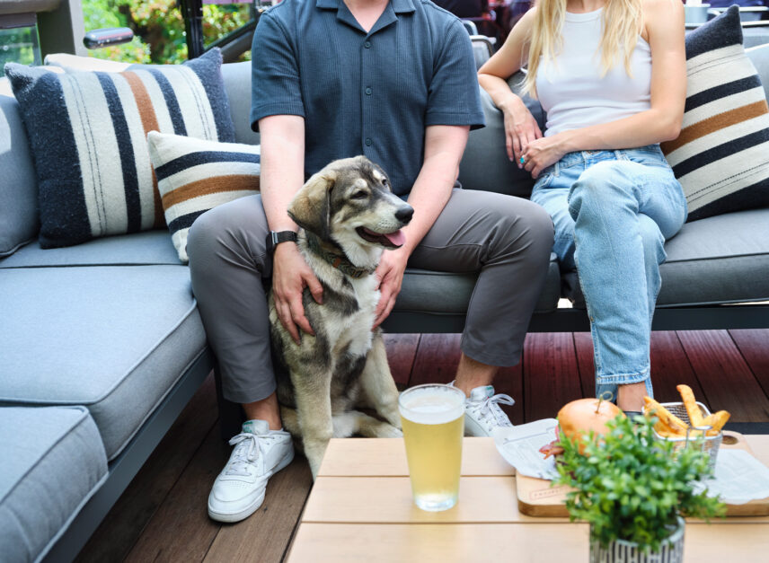 A picture of a dog and a couple sat on the sofas on the patio of Stanley's bar and grill. There is a beer and a burger on the table beside them.