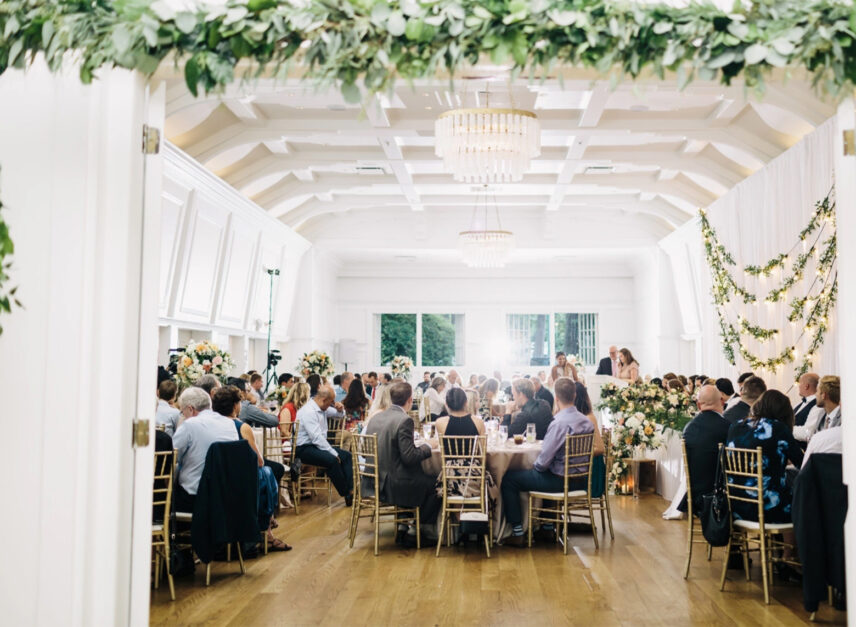The wedding reception in the Stanley Park Pavilion, guests eating at tables while watching people give speeches