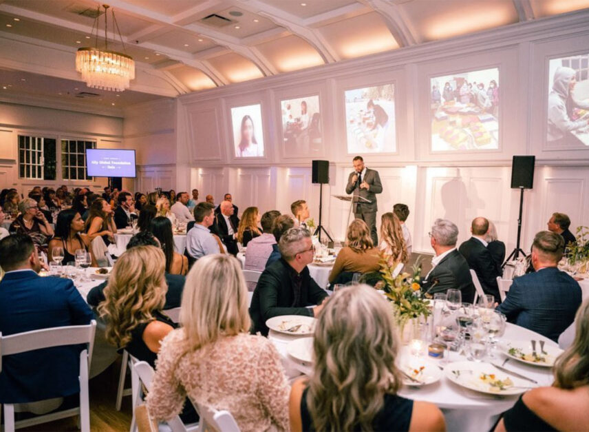 Corporate event, with a man in a suit center stage, speaking with corporate images projected behind him. The audience has finished their food.