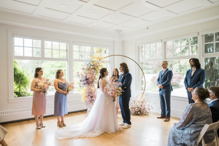 A couple getting married in the Tea Room at the Stanley Park Pavilion. Onlookers look happily as the young couple exchange vows