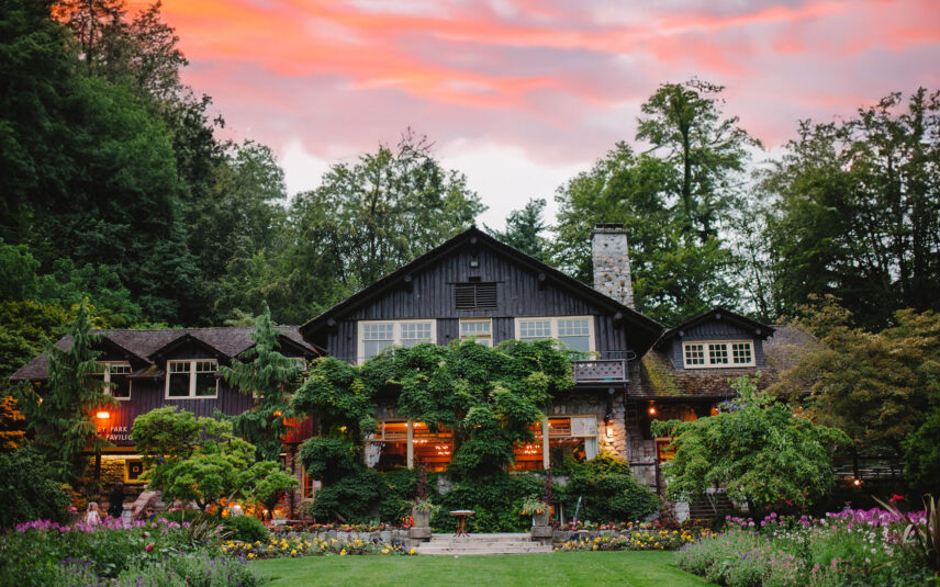 An image of the Stanley Park Pavilion under a red and purple sunset. Colorful flowers flock around, and the warm lights from the Tea Room can be seen.