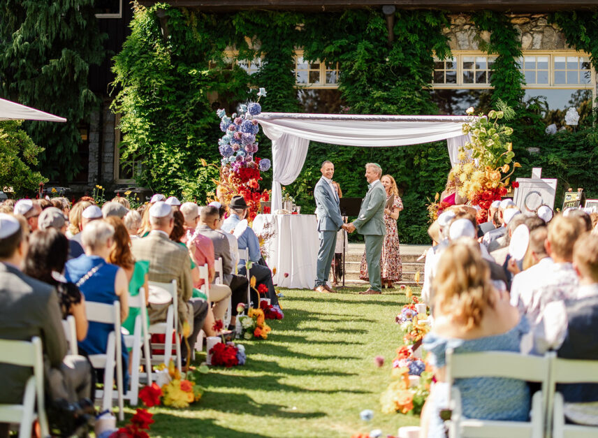A colorful LGBT+ wedding held in front of the Stanley Park Pavilion. Guests are sat in rows, watching the happy couple on a sunny day.