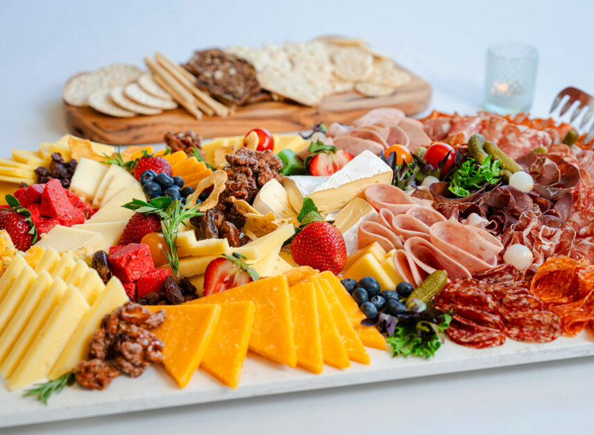 A cheese and meat platter, with assorted fruit. In the background, a board of crackers can be seen.