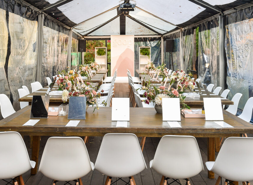 A tent is set up for a corporate event, flowers line the tables and speaker splaying music can be seen in the background.